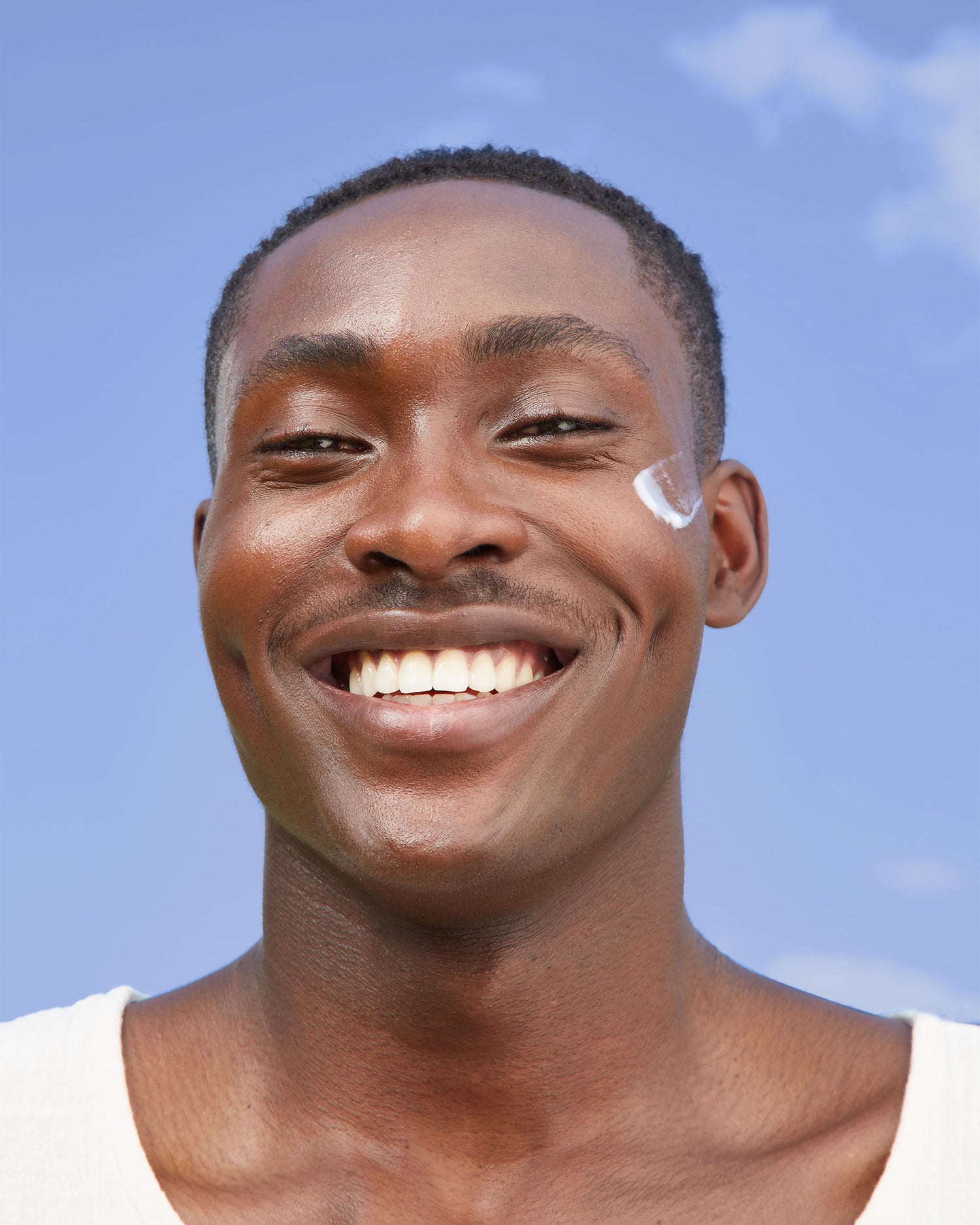 Photo of African American model wearing smear of Michal Morrison Genesis BSTEM6 Molecular Serum on one cheek smiling at camera in front of a bright blue sky background.