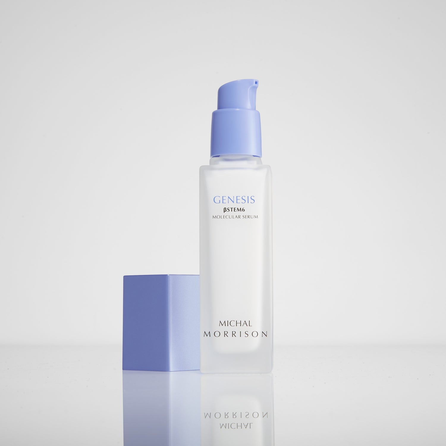 Photo of Michal Morrison Genesis BSTEM6 Molecular Serum in its packaging sitting on gray surface with a white background. The bottle shows a clear rectangular body with a periwinkle pump on top, and a rectangular cap is sitting to its left